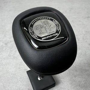 Könnte beinhalten: Schwarzer Lederschaltknauf mit einem Chrom- und schwarzen Emblem. Das Emblem zeigt die Wörter "AFFALTERBACH" und "AMG" mit einem Baum und anderen Designelementen. Der Schaltknauf ist auf einem schwarzen Ständer montiert.