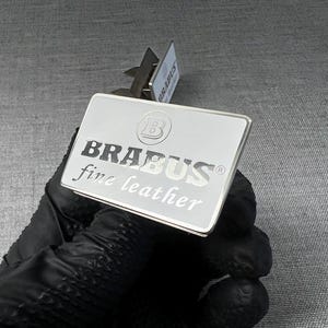 May include: A rectangular silver metal plate with the word "BRABUS" in bold, black capital letters, and "fine leather" in smaller, italicized letters. The plate is held by a gloved hand.