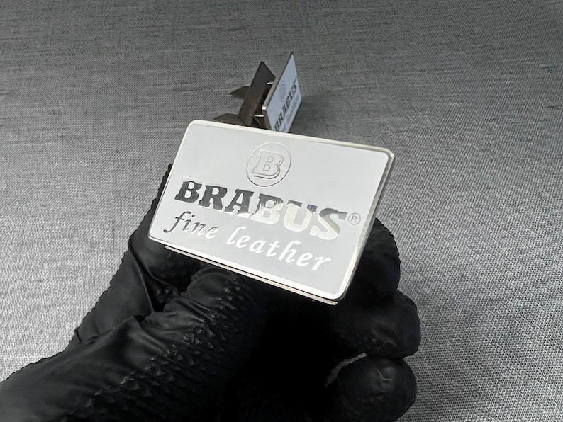 May include: A rectangular silver metal plate with the word "BRABUS" in bold, black capital letters, and "fine leather" in smaller, italicized letters. The plate is held by a gloved hand.