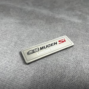 以下が含まれることがあります： 黒、赤、白で「MUGEN Si Limited Edition」の文字が入った長方形のシルバーバッジ。 バッジはつや消し金属仕上げで、グレーのテクスチャード加工された背景に配置されています。