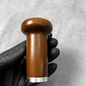 May include: A wooden bottle stopper with a rounded top and a cylindrical body. The wood has a rich, brown color and a visible grain. A silver-colored metal band encircles the base of the stopper.