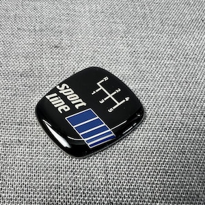 Może przedstawiać: Czarny emblemat Sport Line z wzorem zmiany biegów i niebieskimi paskami. Emblemat ma numery od 1 do 5 i R dla biegu wstecznego. Tekst "Sport Line" jest w kolorze białym.