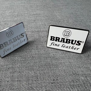 May include: Two rectangular metal and white leather accessories with the text "BRABUS fine leather". One is silver with engraved text, the other is white with black text and a black border. The accessories have the "B" logo.