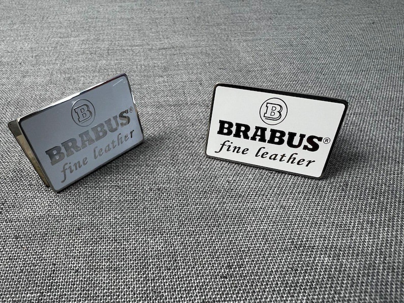 May include: Two rectangular metal and white leather accessories with the text "BRABUS fine leather". One is silver with engraved text, the other is white with black text and a black border. The accessories have the "B" logo.