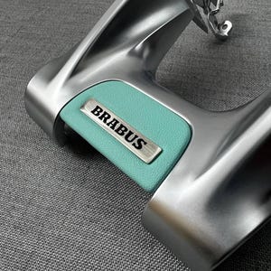 Könnte beinhalten: Nahaufnahme eines silberfarbenen Autoteils mit einem türkisblauen Einsatz. Der Einsatz hat ein silbernes Metallschild mit dem Wort "BRABUS" in schwarzer Schrift. Das Teil hat ein gebogenes Design.