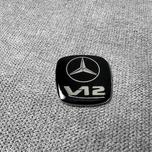 Op de afbeelding: Zwart, vierkant embleem met een zilveren Mercedes-Benz logo en de tekst "V12". Het embleem ligt op een grijze, getextureerde ondergrond. Het Mercedes-Benz logo is een driepuntige ster in een cirkel.