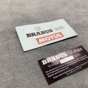 Può includere: Due adesivi in bianco e nero con il testo "BRABUS V-max" e "BRABUS MOTUL". Gli adesivi sono su una superficie grigia.
