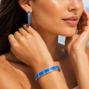 Peut inclure: Boucles d'oreilles créoles argentées et bracelet assorti avec incrustations rectangulaires en opale bleue. Les bijoux sont présentés sur le bras et l'oreille d'une personne, mettant en valeur le bleu vibrant et le design élégant.