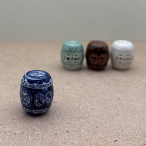 May include: A blue and white ceramic miniature barrel-shaped stool with detailed patterns. Three other miniature stools in green, brown, and white are slightly out of focus in the background. The stools have a decorative design.