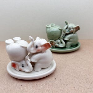 Puede incluir: Dos juegos de figuritas de elefantes de cerámica. Uno es blanco con detalles rosas y rojos, y el otro es verde. Los elefantes están sobre pequeñas bases redondas. Probablemente saleros y pimenteros.