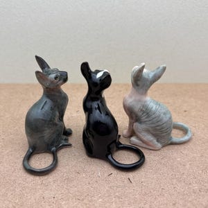 Puede incluir: Tres figuritas de cer&aacute;mica de gatos, una gris, una negra y una gris y blanca, todas mirando hacia arriba.