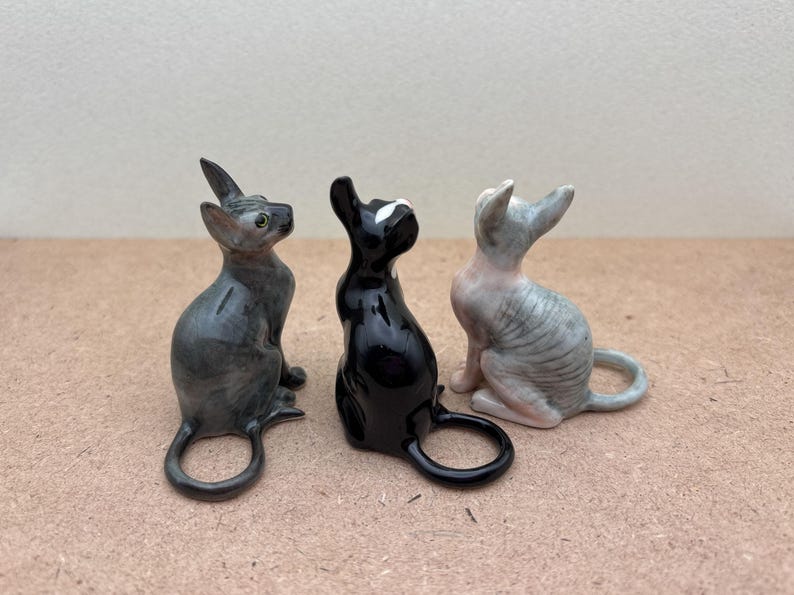 Puede incluir: Tres figuritas de cer&aacute;mica de gatos, una gris, una negra y una gris y blanca, todas mirando hacia arriba.