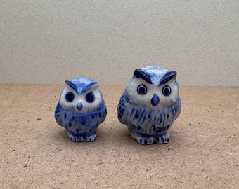 Könnte beinhalten: Zwei blaue und weiße Keramik-Eulenfiguren. Die größere Eule steht mit ausgebreiteten Flügeln. Die kleinere Eule sitzt mit eingeklappten Flügeln.