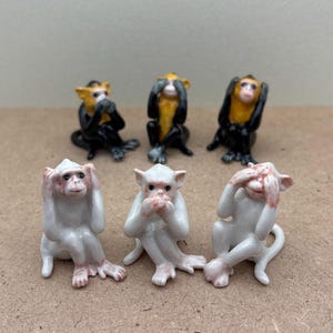 May include: Five porcelain monkey figurines, each with a different color and pose. One monkey is black, one is yellow, one is white, and two are white with pink accents. The monkeys are all covering their eyes, ears, or mouth.