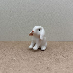 May include: A small white ceramic rabbit figurine with black eyes and a pink nose. The rabbit is sitting on a brown surface.