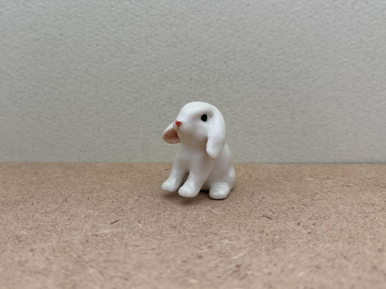 May include: A small white ceramic rabbit figurine with black eyes and a pink nose. The rabbit is sitting on a brown surface.