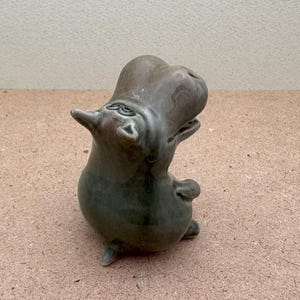 K&ouml;nnte beinhalten: Eine Keramikfigur eines gr&uuml;nen Flusspferdes mit nach hinten geneigtem Kopf und offenem Maul. Das Flusspferd steht auf einer hellbraunen Oberfl&auml;che.