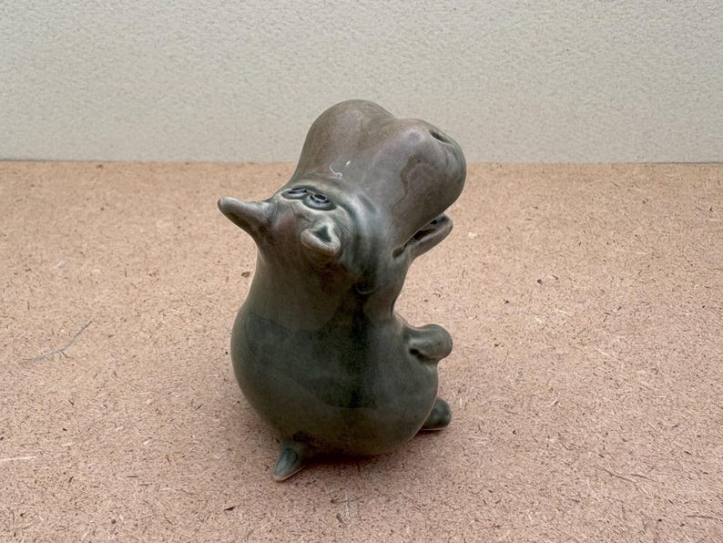K&ouml;nnte beinhalten: Eine Keramikfigur eines gr&uuml;nen Flusspferdes mit nach hinten geneigtem Kopf und offenem Maul. Das Flusspferd steht auf einer hellbraunen Oberfl&auml;che.