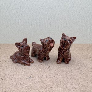Puede incluir: Tres figuritas de cer&aacute;mica marr&oacute;n de perros Yorkshire Terrier. Los perros est&aacute;n en diferentes poses, uno est&aacute; sentado, otro est&aacute; de pie y otro est&aacute; acostado.