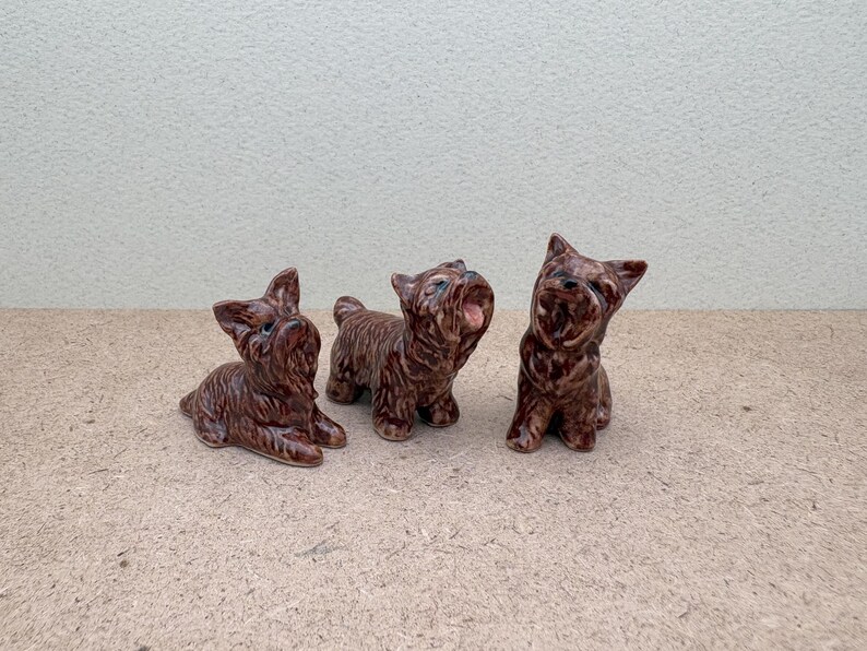 Puede incluir: Tres figuritas de cer&aacute;mica marr&oacute;n de perros Yorkshire Terrier. Los perros est&aacute;n en diferentes poses, uno est&aacute; sentado, otro est&aacute; de pie y otro est&aacute; acostado.