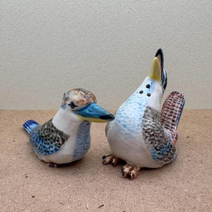 May include: A pair of ceramic salt and pepper shakers shaped like kookaburras. The shakers are painted in shades of blue, white and brown, with realistic details. The kookaburra on the left is facing the right, while the kookaburra on the right is facing the left.