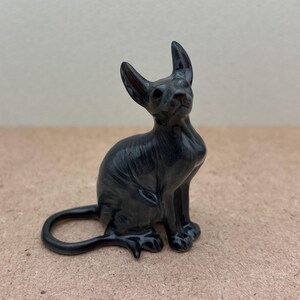 K&ouml;nnte beinhalten: Eine schwarze Keramikfigur eines haarlosen Katers, der mit seinem Schwanz um seine Beine gewickelt sitzt.