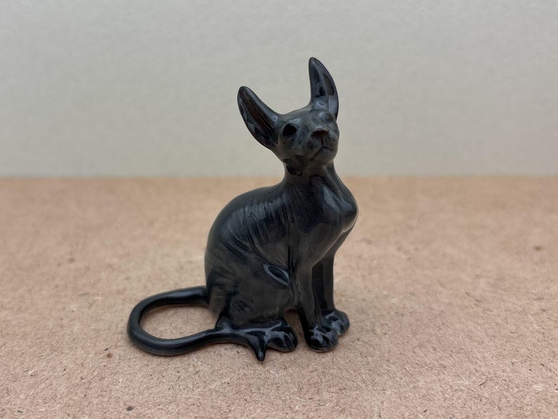 K&ouml;nnte beinhalten: Eine schwarze Keramikfigur eines haarlosen Katers, der mit seinem Schwanz um seine Beine gewickelt sitzt.