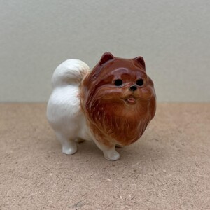 May include: A small ceramic figurine of a brown and white Pomeranian dog. The dog is standing on all four paws and has a fluffy tail.