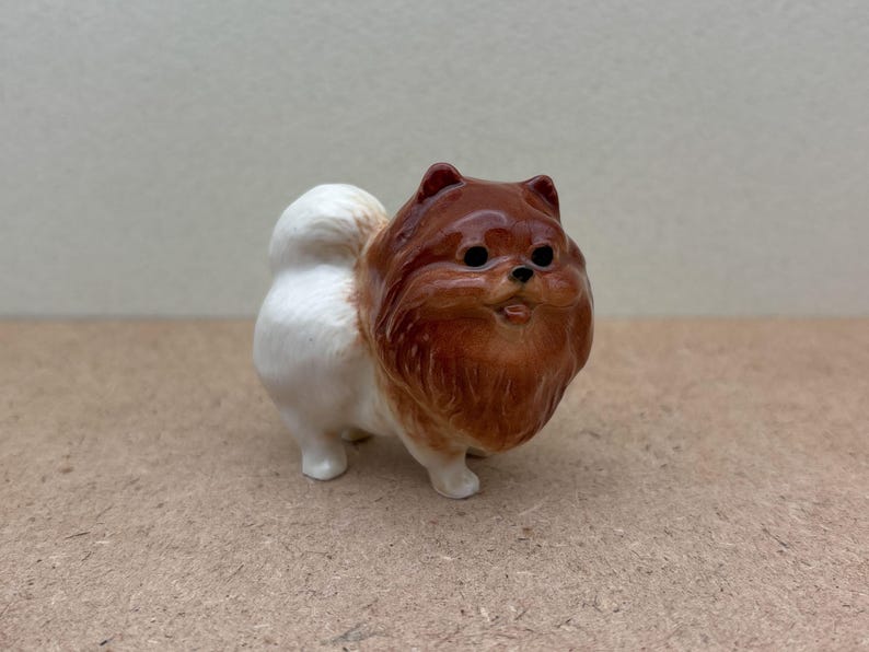 May include: A small ceramic figurine of a brown and white Pomeranian dog. The dog is standing on all four paws and has a fluffy tail.