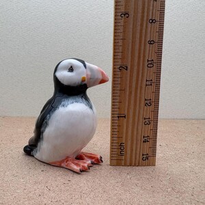 May include: A ceramic figurine of a black and white puffin with an orange beak. The figurine is about 2 inches tall and is standing next to a ruler.