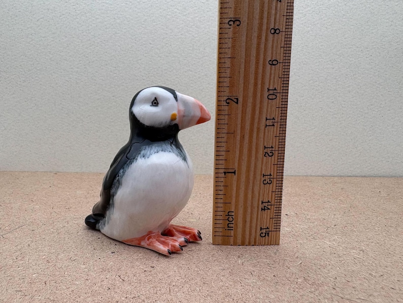 May include: A ceramic figurine of a black and white puffin with an orange beak. The figurine is about 2 inches tall and is standing next to a ruler.