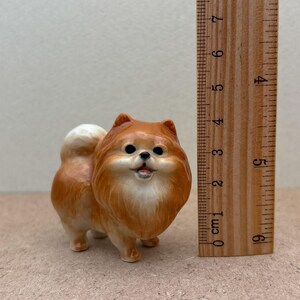 May include: A small ceramic figurine of a brown and white Pomeranian dog. The figurine is about 3 inches tall and is standing on a brown surface.