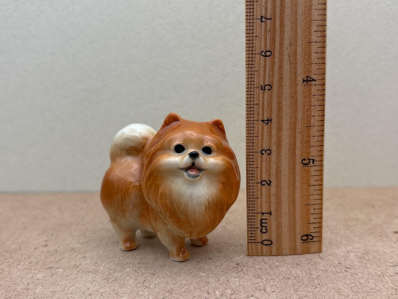 May include: A small ceramic figurine of a brown and white Pomeranian dog. The figurine is about 3 inches tall and is standing on a brown surface.