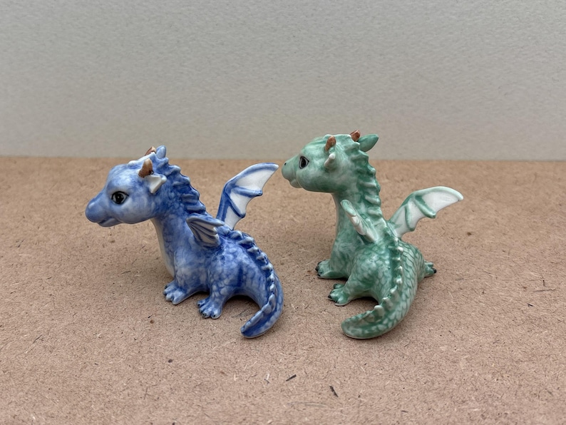 May include: Two small ceramic dragon figurines. One is blue with white wings and the other is green with white wings.