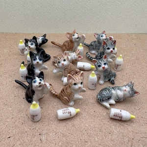 May include: A collection of 12 ceramic kitten figurines, each with a different pose and colour pattern. The kittens are all holding miniature milk bottles.