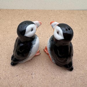May include: Two ceramic salt and pepper shakers shaped like puffins. The puffins are black and white with orange beaks and feet. They are facing each other.