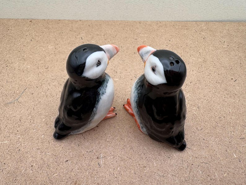 May include: Two ceramic salt and pepper shakers shaped like puffins. The puffins are black and white with orange beaks and feet. They are facing each other.