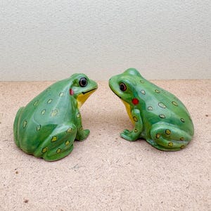 Puede incluir: Dos figuras de ranas de cer&aacute;mica verdes. Las ranas tienen vientre amarillo, una mancha roja en la mejilla y ojos oscuros. Est&aacute;n decoradas con peque&ntilde;as manchas verde oscuro y se miran.