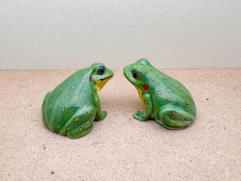 Puede incluir: Dos figuras de ranas de cer&aacute;mica verdes. Las ranas tienen vientre amarillo, una mancha roja en la mejilla y ojos oscuros. Est&aacute;n decoradas con peque&ntilde;as manchas verde oscuro y se miran.