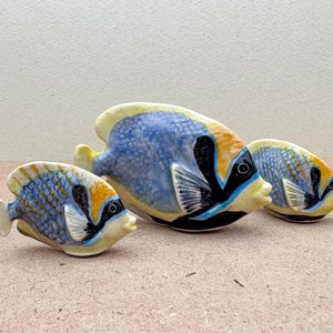 Może przedstawiać: Trzy ceramiczne figurki ryb w kolorze niebieskim, żółtym i czarnym. Największa ryba znajduje się w środku, z mniejszą rybą po obu stronach. Ryby są skierowane w prawo.