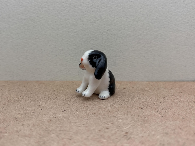 May include: A small ceramic figurine of a black and white rabbit sitting on a brown surface. The rabbit has a pink nose and black eyes.
