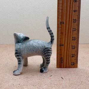 K&ouml;nnte beinhalten: Kleine Katzenfigur aus Keramik in Grau- und Schwarzt&ouml;nen. Die Katze steht mit nach oben geneigtem Kopf und erhobenem Schwanz. Ein Holzlineal liegt neben der Katze, das Zoll anzeigt.