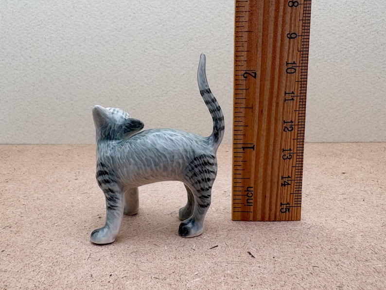 K&ouml;nnte beinhalten: Kleine Katzenfigur aus Keramik in Grau- und Schwarzt&ouml;nen. Die Katze steht mit nach oben geneigtem Kopf und erhobenem Schwanz. Ein Holzlineal liegt neben der Katze, das Zoll anzeigt.