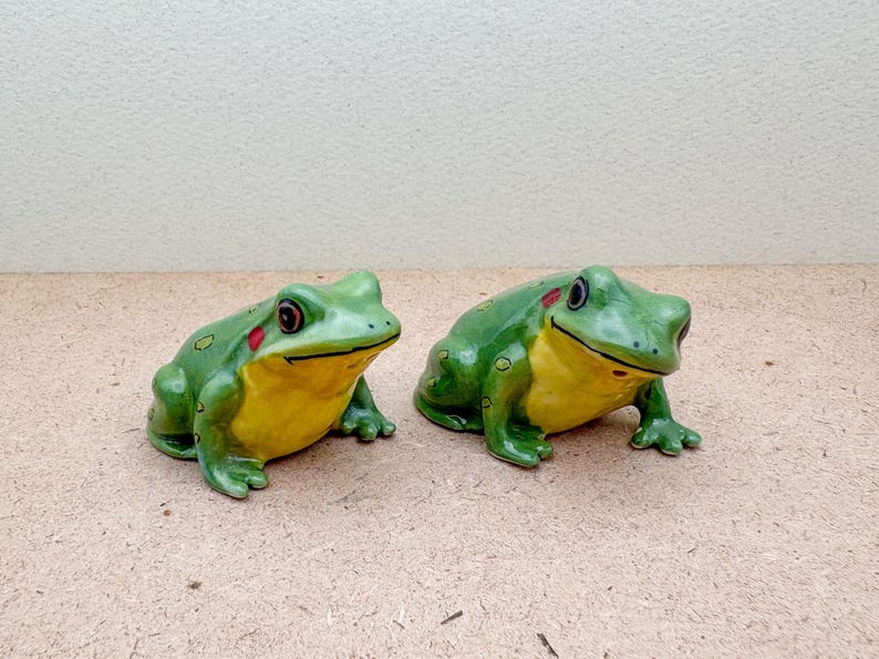 Puede incluir: Dos figuras de rana de cer&aacute;mica. Las ranas son verdes con vientres amarillos, detalles rojos y caras sonrientes. Est&aacute;n colocadas una al lado de la otra sobre una superficie marr&oacute;n claro, mostrando un tema de decoraci&oacute;n de jard&iacute;n caprichoso.