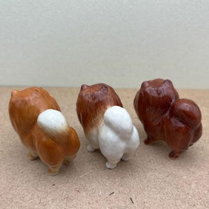 May include: Three ceramic dog figurines, each with a different color fur. The dog on the left has brown and white fur, the middle dog has white and brown fur, and the dog on the right has brown fur.
