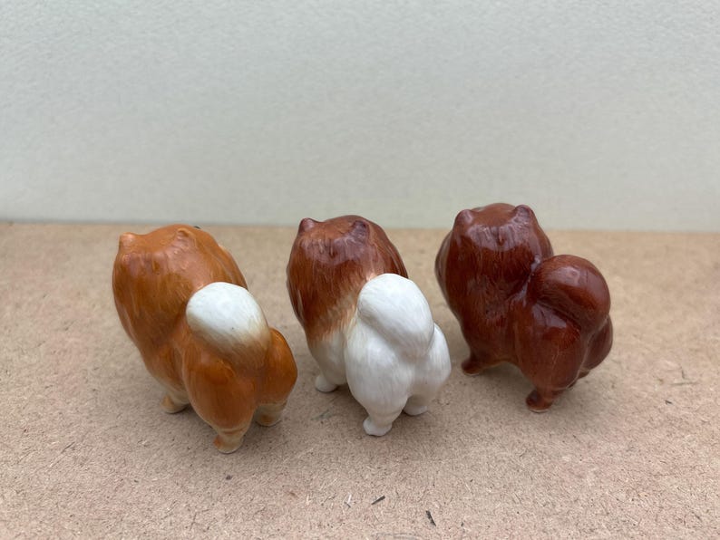 May include: Three ceramic dog figurines, each with a different color fur. The dog on the left has brown and white fur, the middle dog has white and brown fur, and the dog on the right has brown fur.