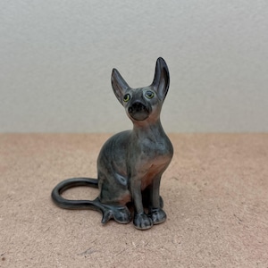 Puede incluir: Una peque&ntilde;a figurita de cer&aacute;mica gris de un gato sin pelo sentado con la cola enroscada alrededor de sus patas. El gato tiene ojos verdes y una nariz gris clara.