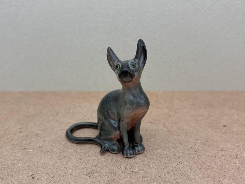 Puede incluir: Una peque&ntilde;a figurita de cer&aacute;mica gris de un gato sin pelo sentado con la cola enroscada alrededor de sus patas. El gato tiene ojos verdes y una nariz gris clara.
