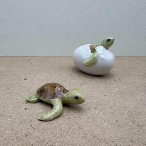 May include: Two ceramic turtle figurines. One is a small, green and brown turtle. The other is a turtle hatching from a white egg. The turtle's head and limbs are green, and the shell is brown.