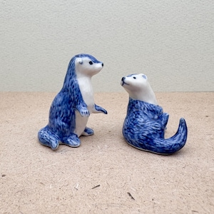 Könnte beinhalten: Zwei blaue und weiße Keramik-Otter-Figuren. Ein Otter steht aufrecht, während der andere mit dem Schwanz um den Körper gewickelt sitzt.